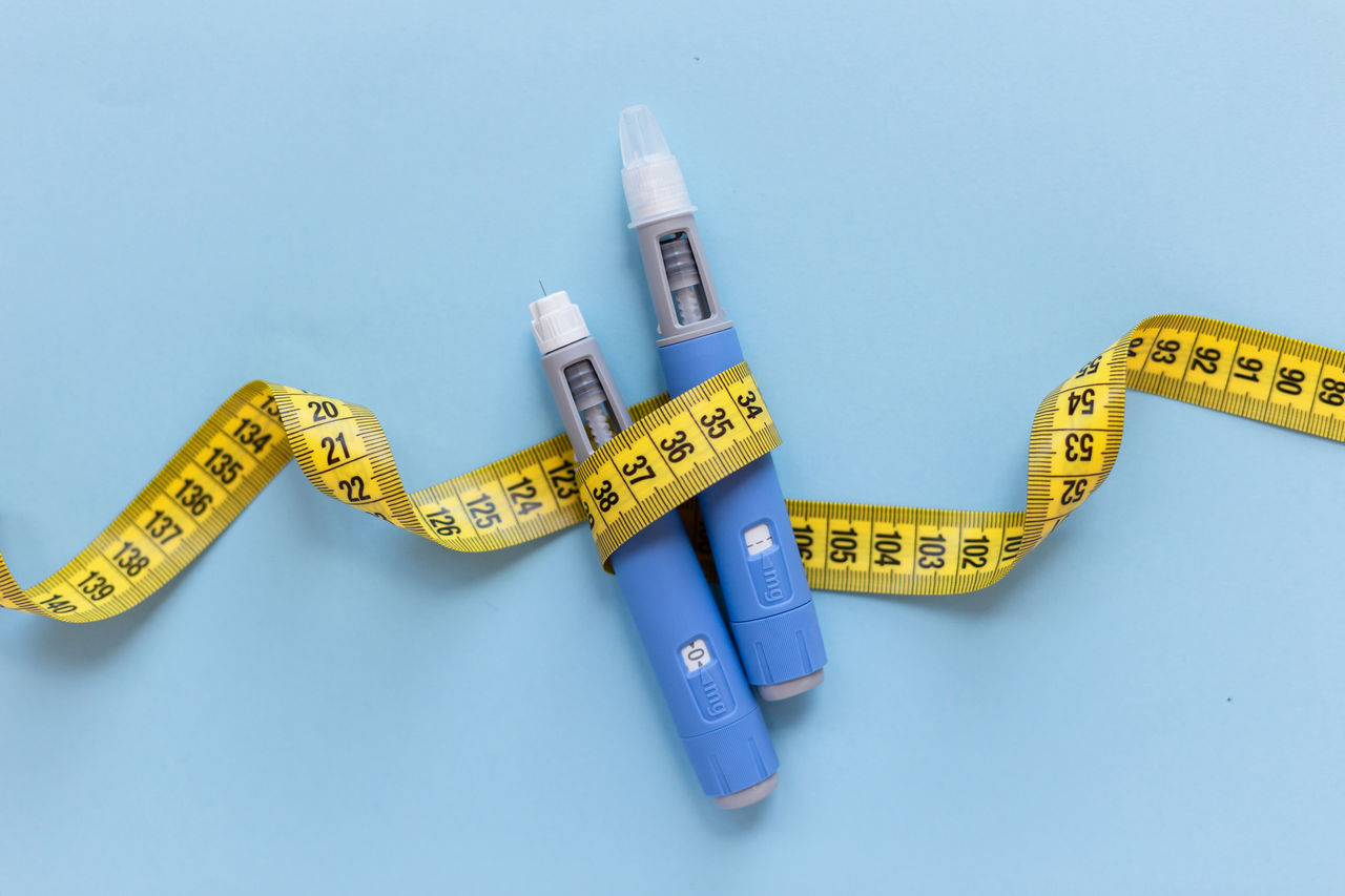 Two weight loss drug injection pens over a blue background wrapped in yellow measuring tape. 