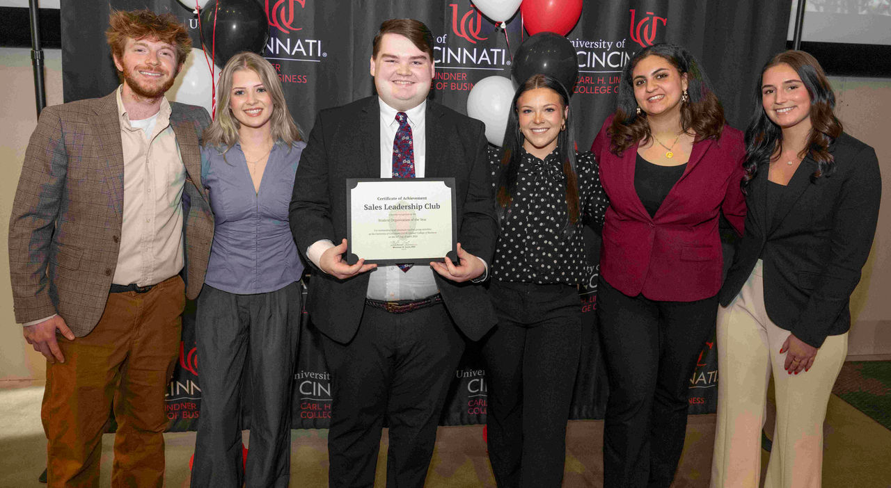 Six students in professional dress stand in front of a black, Lindner-branded banner.