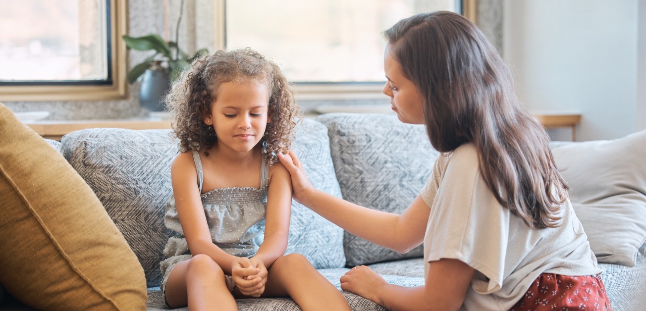 A woman places a hand on the shoulder of a child sitting on a couch