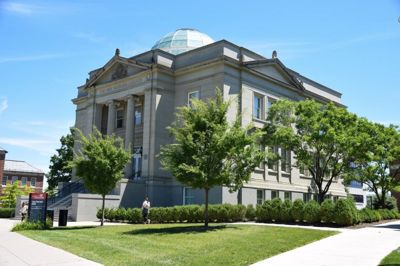 A historic academic building with stone columns and a glass dome, surrounded by trees and walkways on a sunny day.