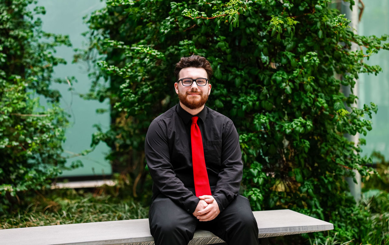 Marcus Elliott, wearing a red tie and black shirt and causal pants, sits on a bench in a garden courtyard at Lindner College of Business.