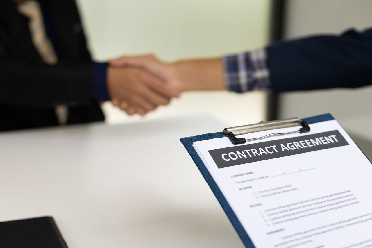 shows two people shaking hands and there is contract agreement on a clipboard in the forefront