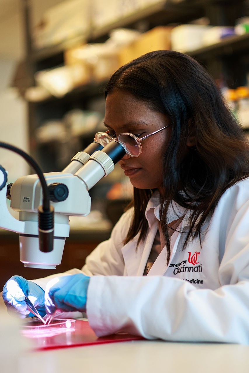 Abhinaya Durairaj in a white lab coat looking nto a microscope
