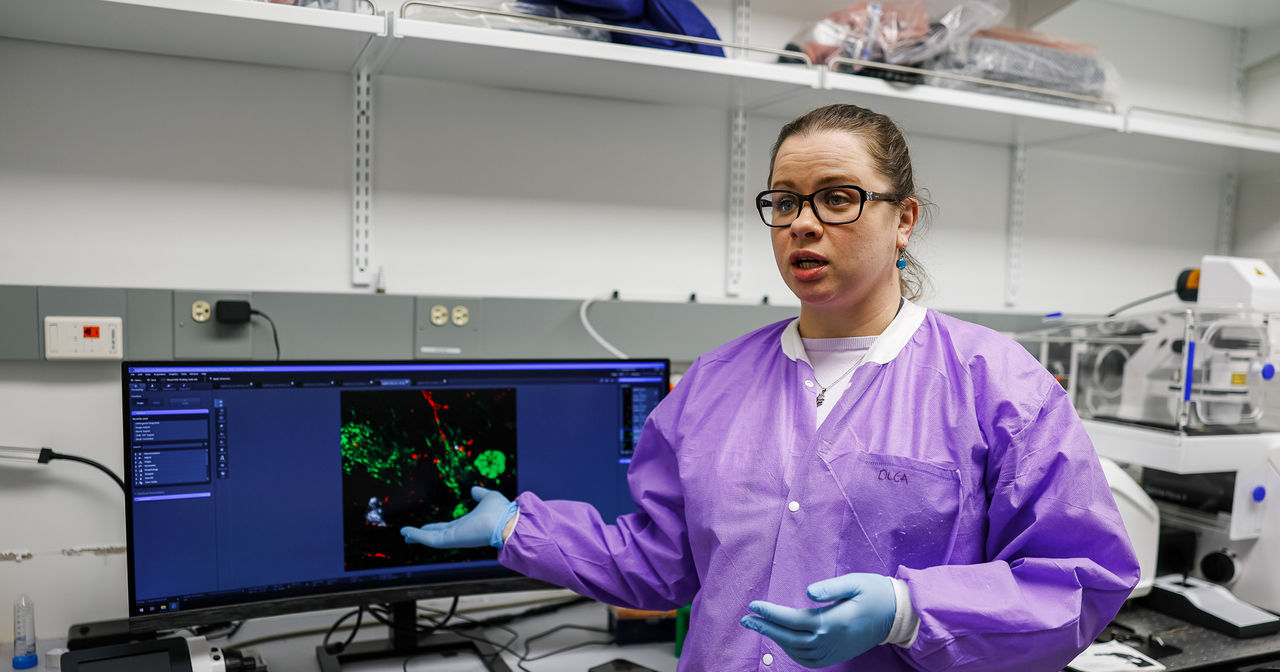 Olga Liaudanskaya dressed in a labcoat and gloves talks about her research in front of a computer showing the function of brain cells.