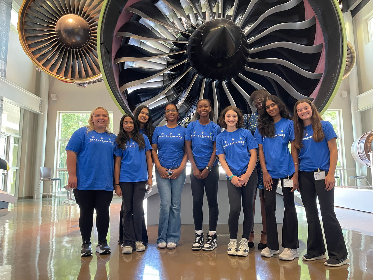 Next Engineers students in matching T-shirts stand in front of a jet engine.