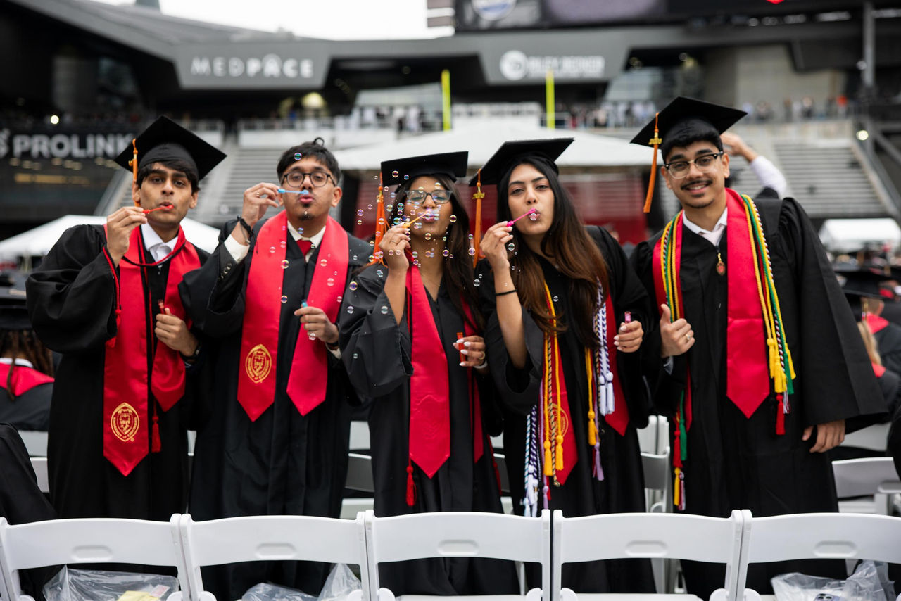 Five students in caps and gowns blow bubbles in front of their seats on the field.