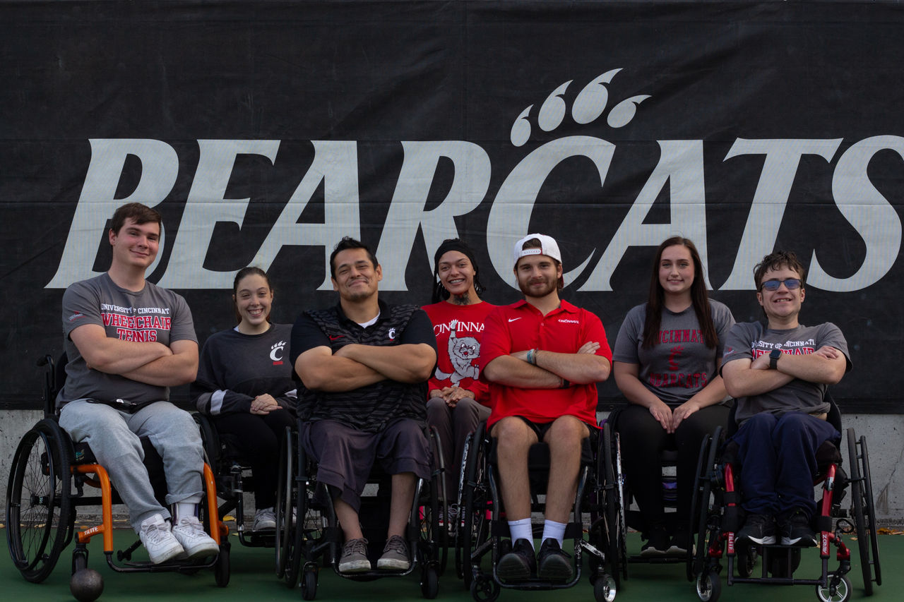 7 members of the UC Adaptive Athletics team line up in front of a Bearcats banner