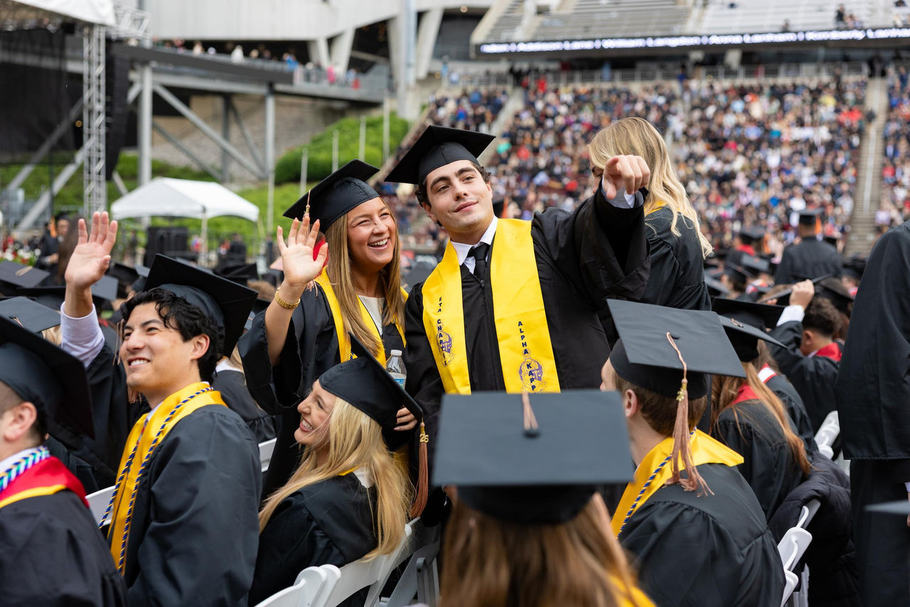 Graduates in caps and gowns wave to supporters in the stands at Nippert Stadium.