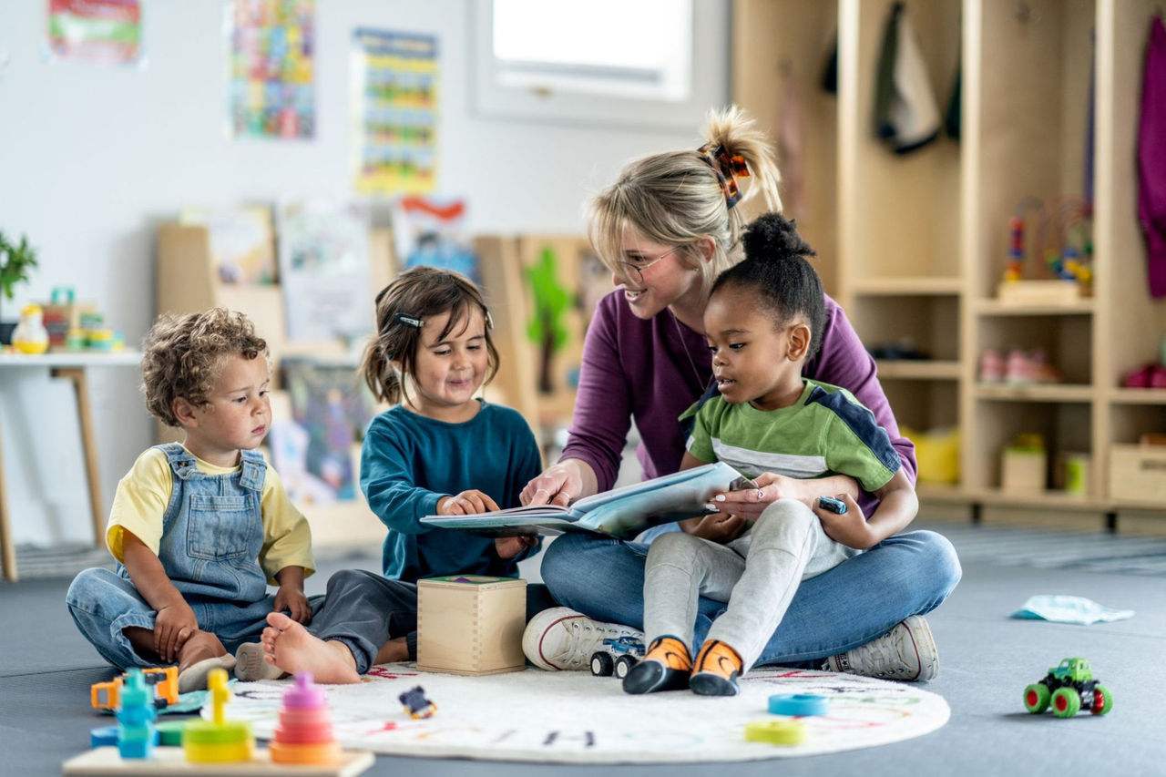 Teaching sitting on a cushion holding one preschool girl and two other preschool child are at her side.  They are reading a book.
