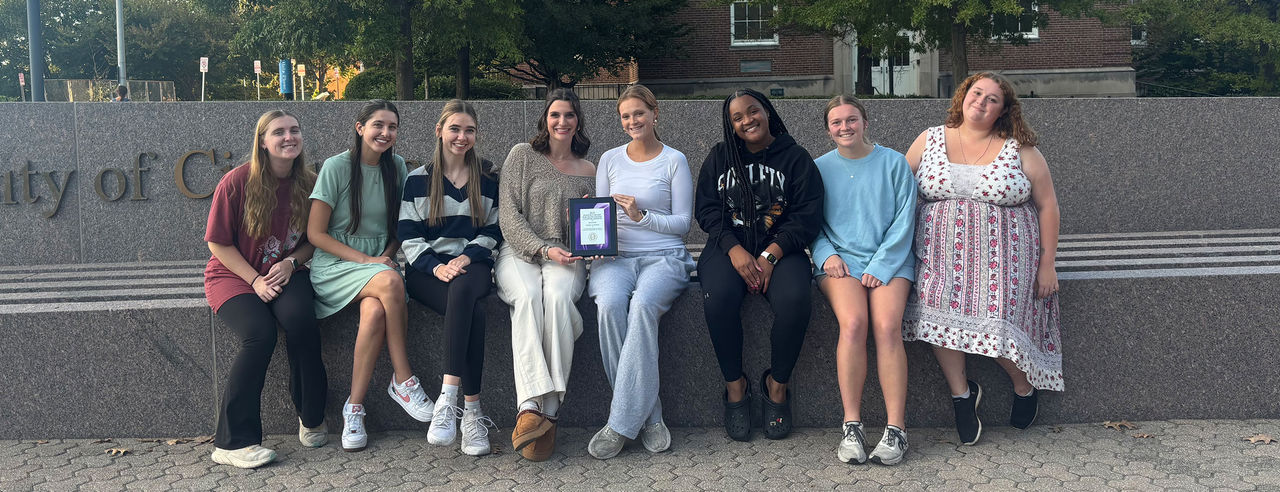 Members of UC's Kappa Delta Pi sit together in front of the University of Cincinnati sign, the middle two people holding an award certificate