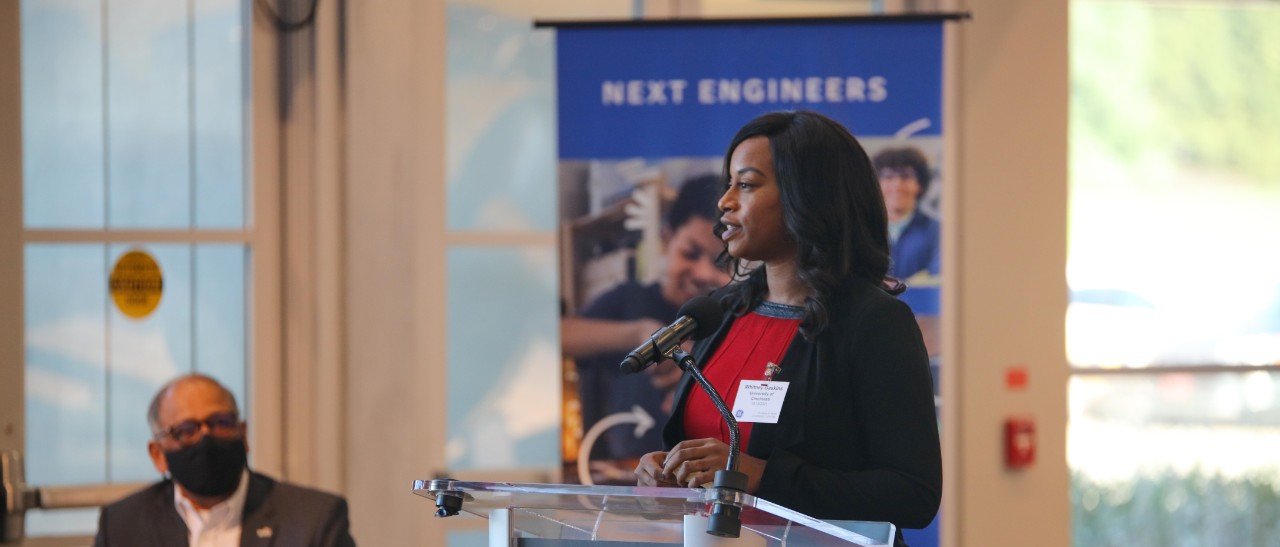 Whitney Gaskins stands at a podium in front of a Next Engineers poster.