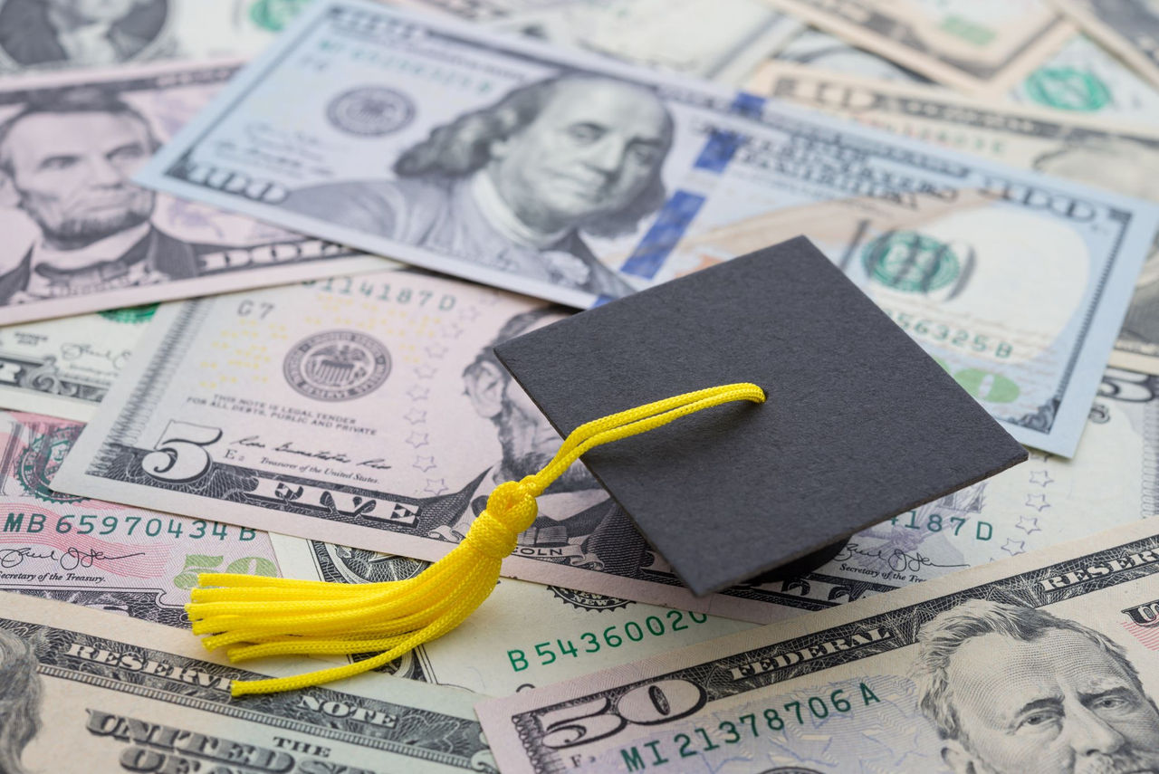printed dollars bills in the background with a graduation hat sitting on top