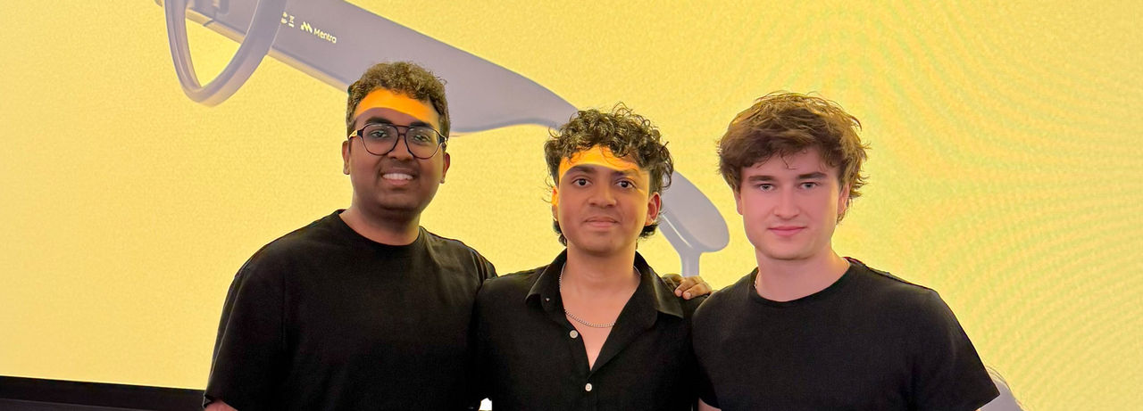 Three people wearing dark shirts stand close together, smiling, in front of a bright yellow backdrop showing an enlarged image of sleek, modern eyeglass frames.