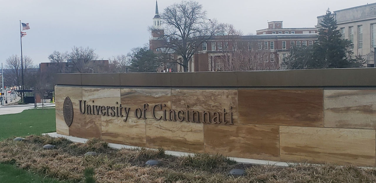 A sign at the university's main gateway showing University of Cincinnati is displayed with campus building in the background.