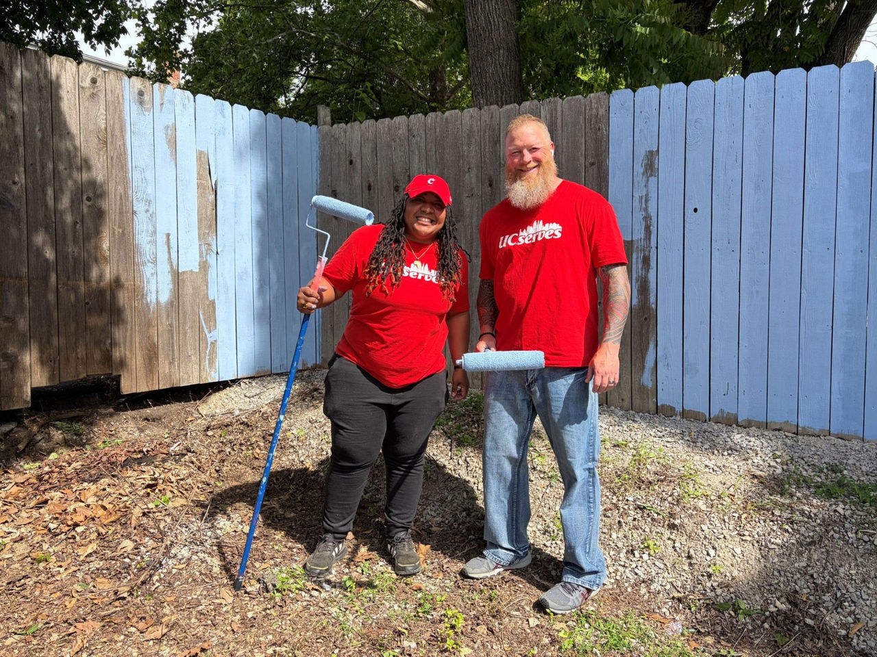 Two UC employees wearing UC Serves shirts hold paint rollers and stand in front of a half painted fence.