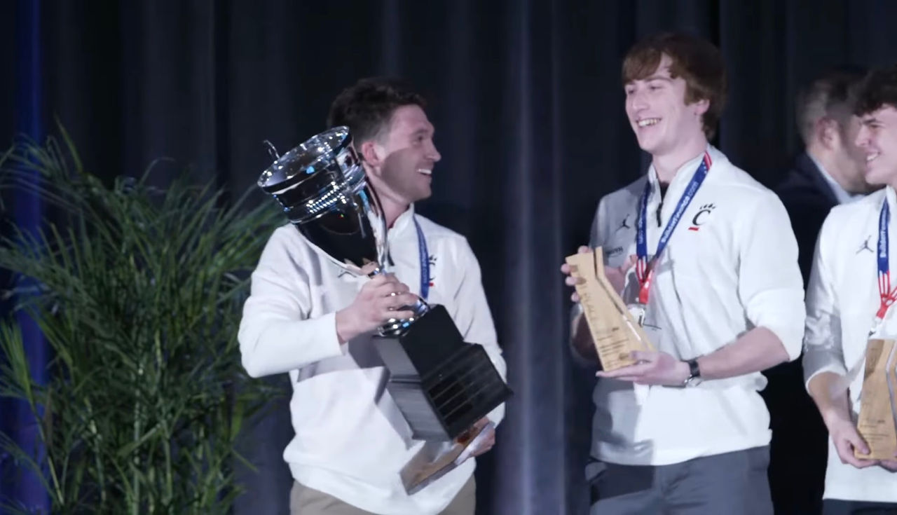 Joe Basti smiles, holding a large silver trophy. Next to him, Daniel Schlegel smiles, holding an award plaque. 