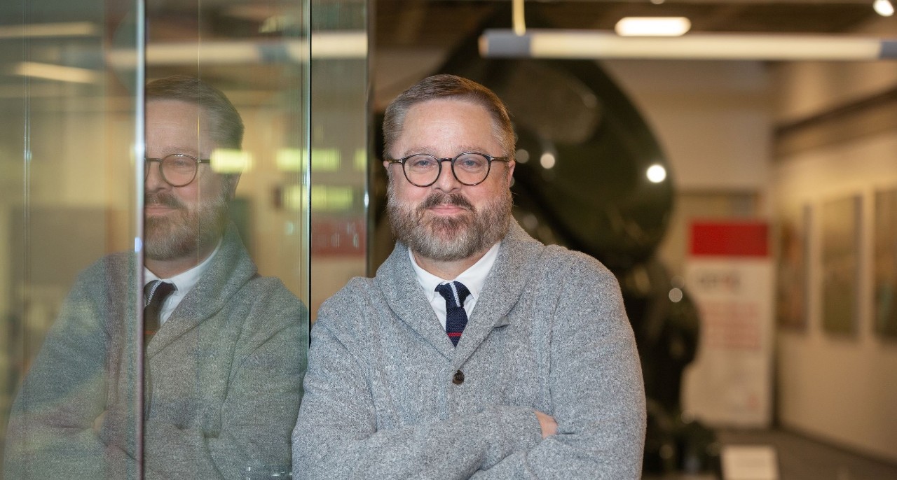 UC Professor Jeffrey Blevins poses next to a glass wall that reflects his image.