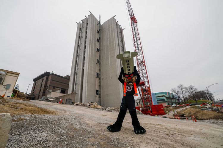The UC Bearcat mascot holds up a model of Crosley Tower in front of the demolition site of Crosley Tower. 