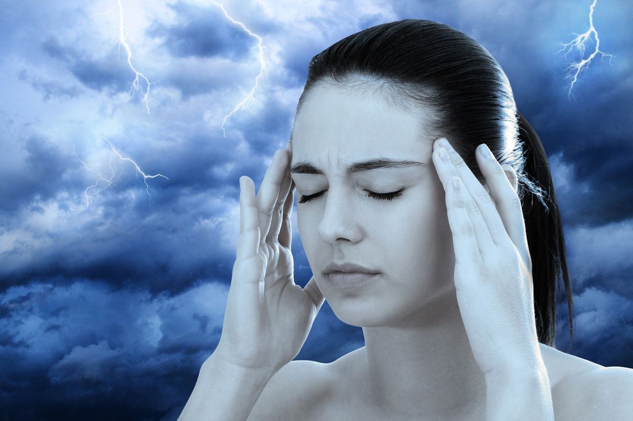 Woman holds her fingers to her temples with lightning and storm clouds in the background, illustrating the connection between weather and migraine headaches