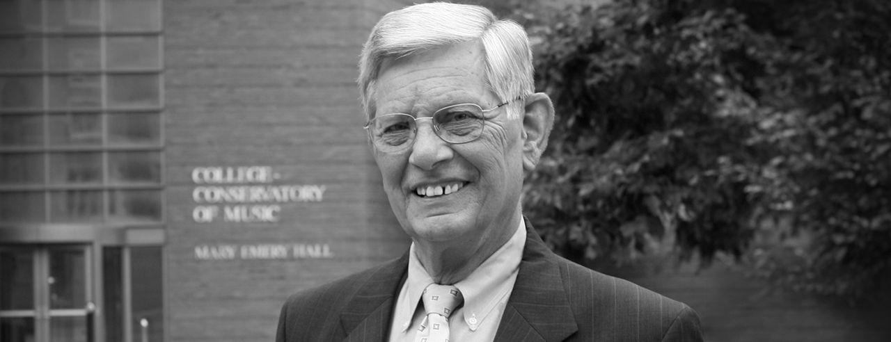 A black and white photograph of CCM Associate Dean Emeritus Warren George standing in front of the main entrance to Mary Emery Hall and the CCM Atrium.