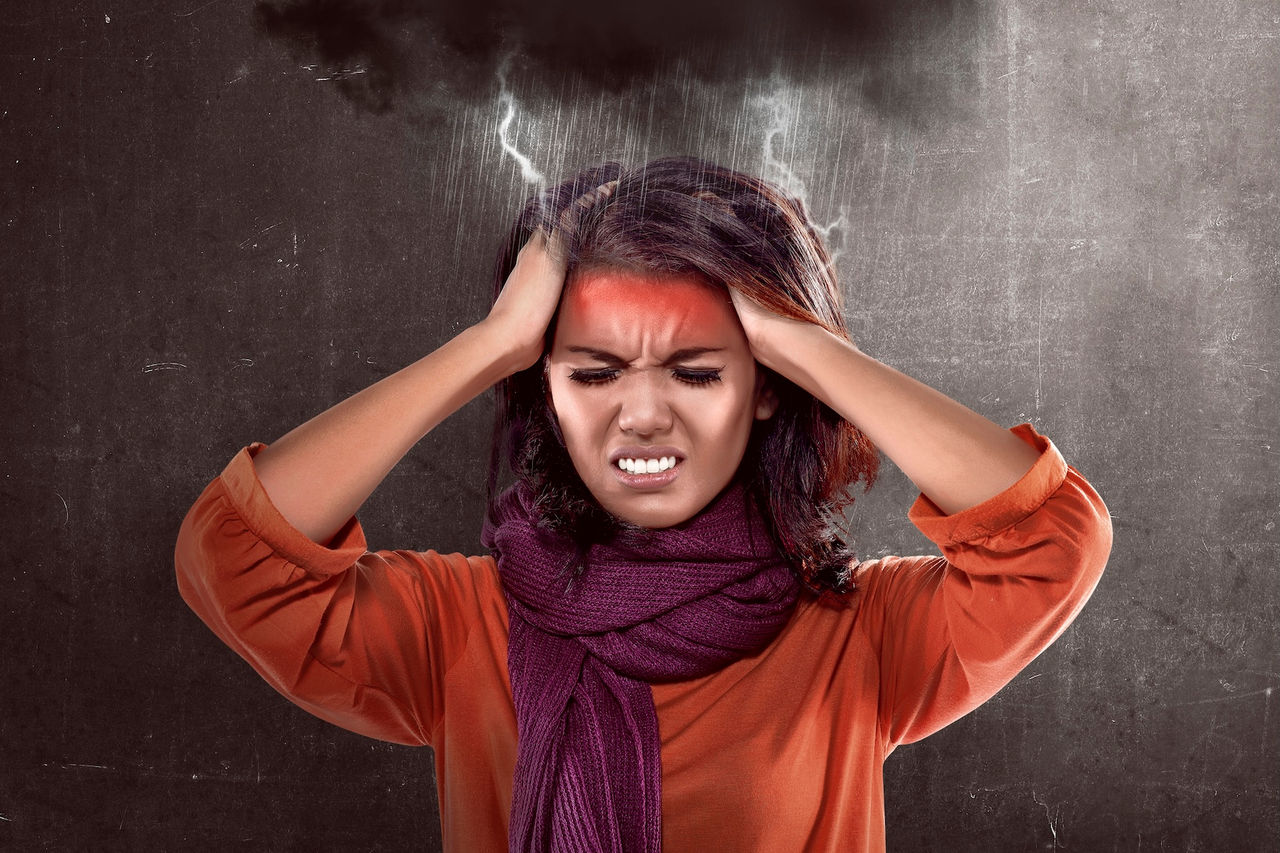 Woman with a scarf around her neck holds her hands to the sides of her head in pain while lightning strikes above her 