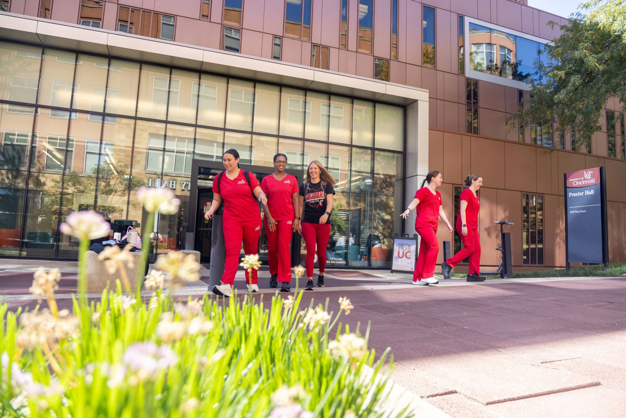 Several nursing students dressed in scrubs,are walking in front of the College of Nursing building Procter Hall.