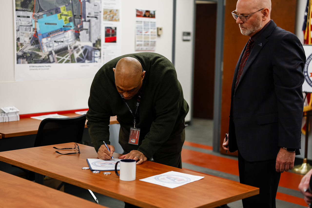 A man leans over a table and signs a piece of paper. A second man looks on.