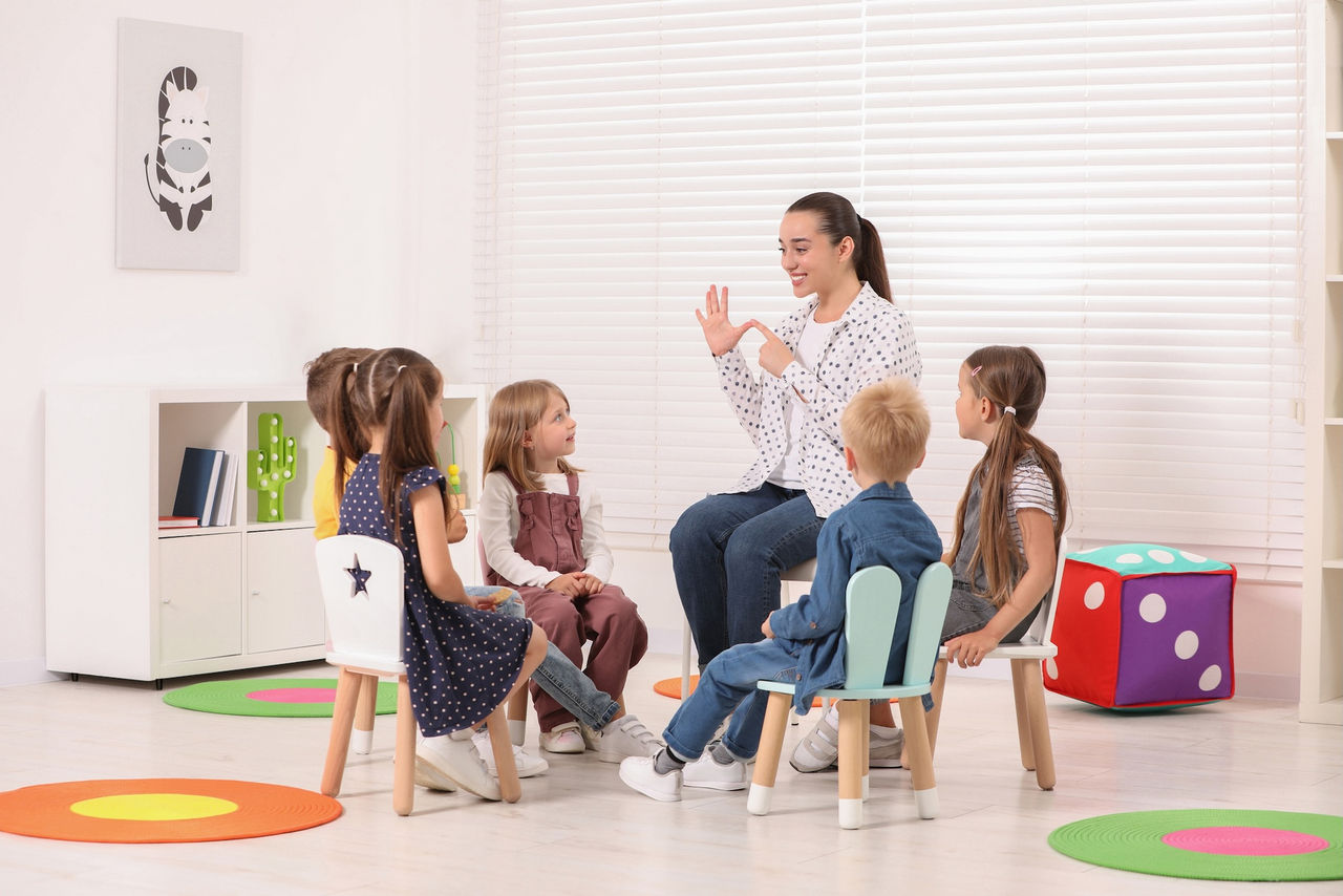 A teacher and preschool students sit on chairs in a circle in a classroom setting and learn numbers and counting 