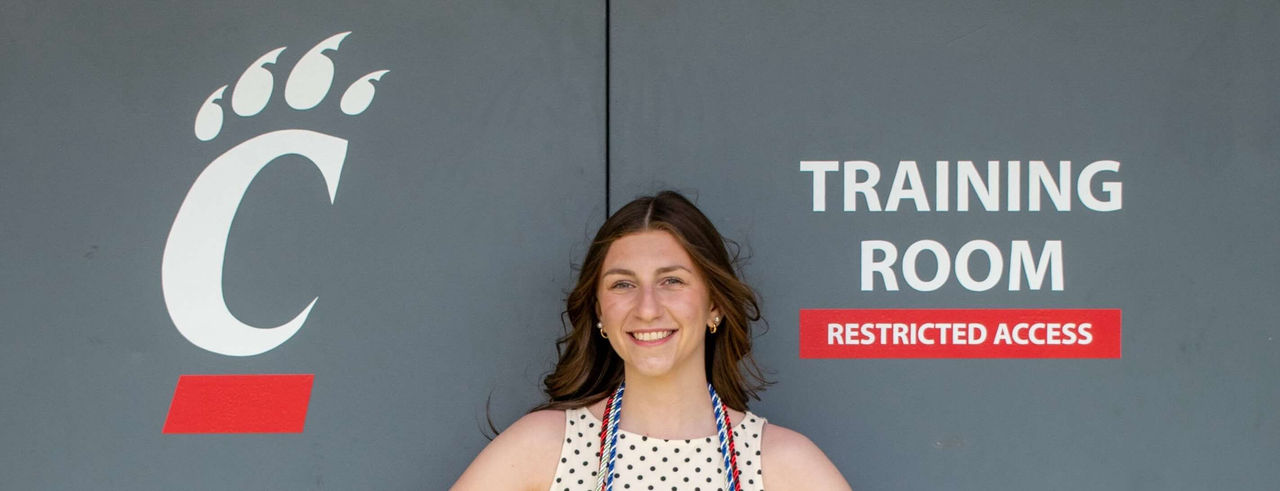 Maddy Dixon stands with her graduation cords in front of the University of Cincinnati training room 