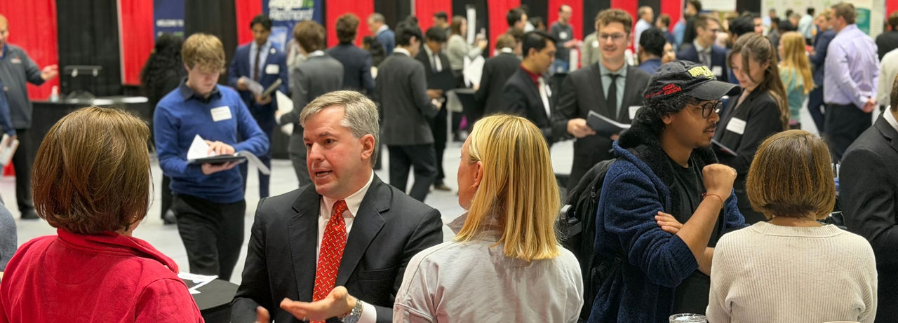 Crowded indoor career fair with students and recruiters in business attire talking in small groups; in the foreground, a man in a suit with a red tie speaks with attendees while others hold resumes and folders, with red and black booth curtains in the background.