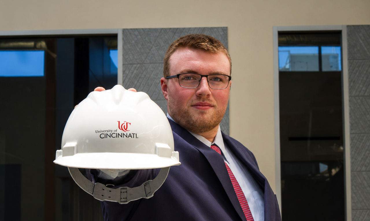 Jeremy Ackley in a suit and tie holds up a hardhat with the UC logo on it.