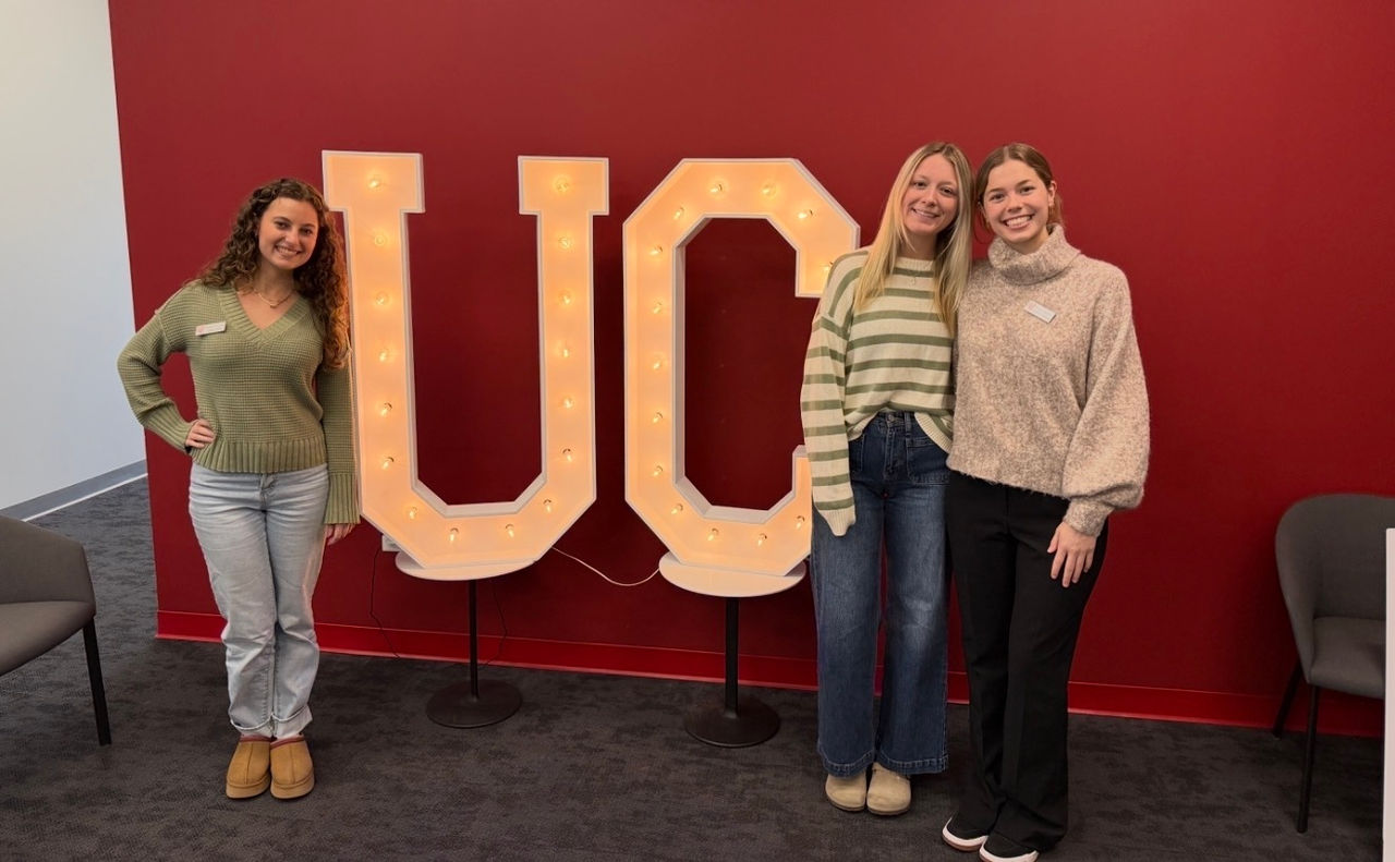 Three graduate students stand next to large, light-up UC letters