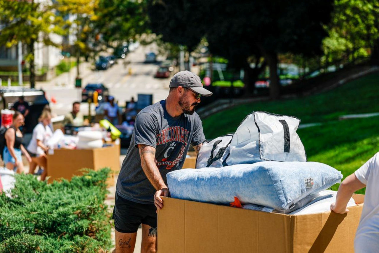 UC Dad is shown pushing a load of items toward a college dorm.  Lots of people behind him in line with dorm essentials.