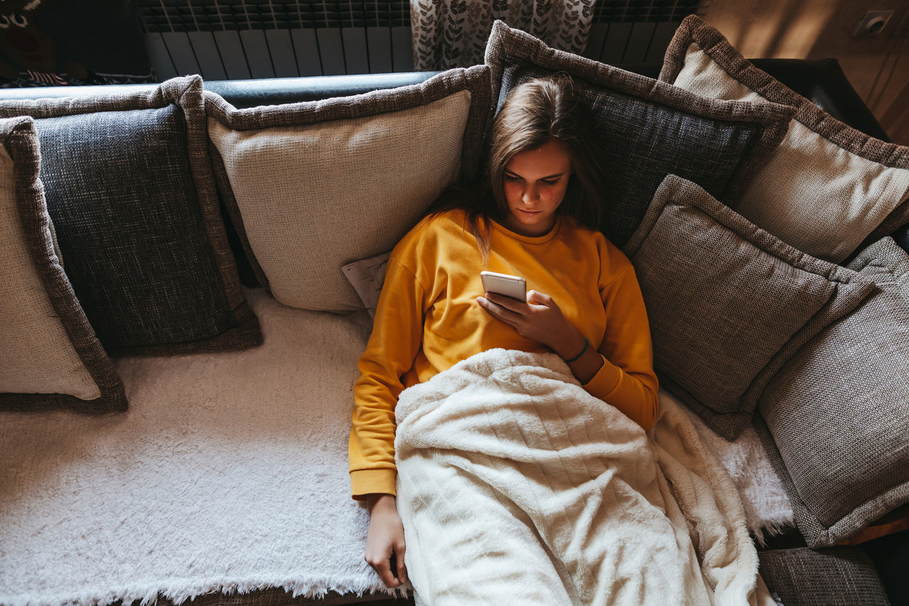 A person in a blanket on the couch scrolls on her phone.