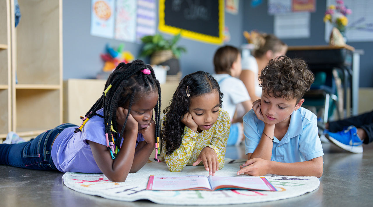Children lie on the floor reading a book in a classroom.