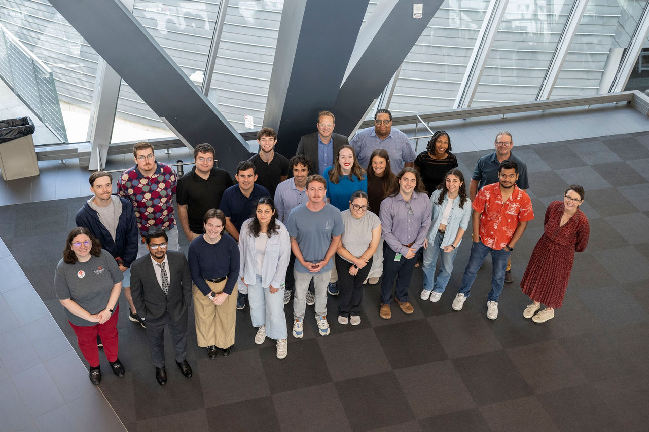 Overhead group photo of approximately 20 people gathered in a bright, modern atrium with large glass windows and exposed steel support beams. The group stands on a dark tiled floor and looks up toward the camera. The space is filled with natural light, and the architectural features of the building create a contemporary, open setting.