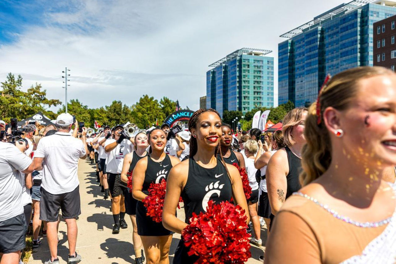 Half a dozen cheerleaders lined up with pompoms and dressed in UC swag to cheer on the Bearcat Football team at a tailgate gathering. Behind them is the UC Marching Band