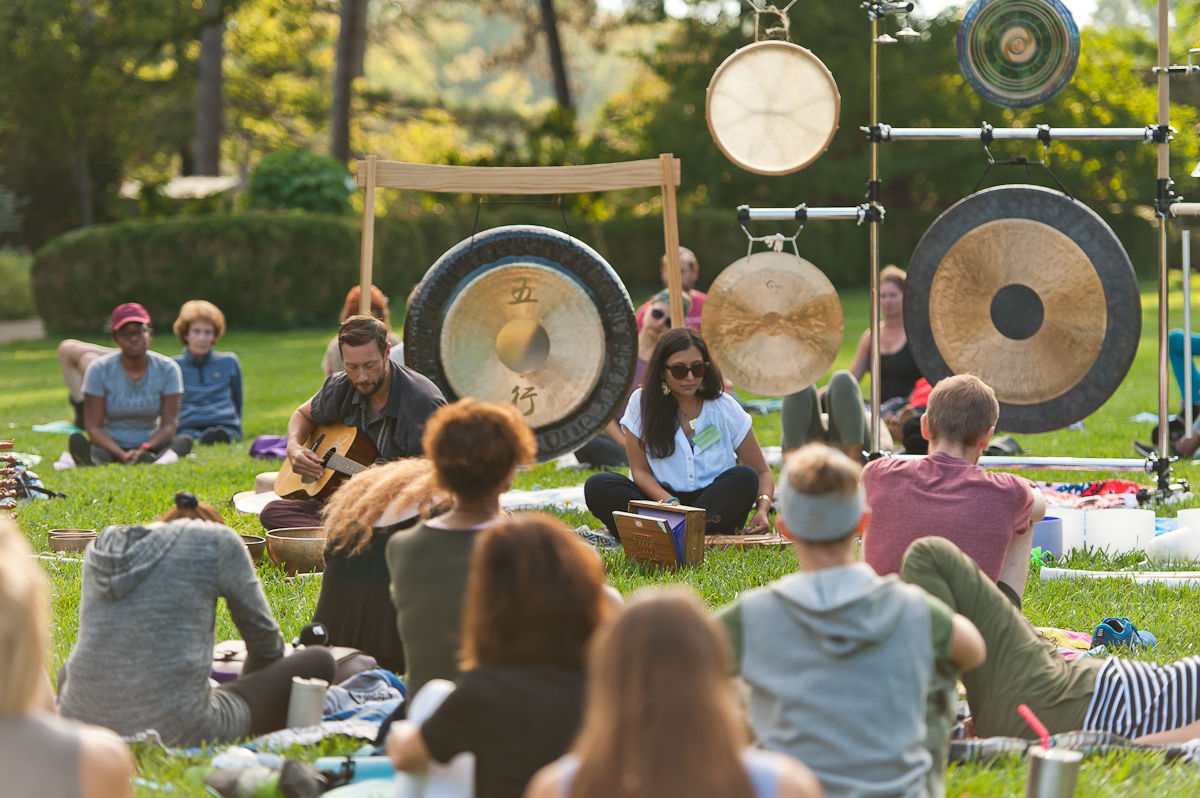 People sitting in the grass in a park. One individual is playing guitar. There are gongs in the background.