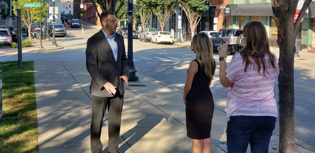 Ben Feldmeyer, PhD, is standing in a Cincinnati park area being interviewed by a WCPO reporter while a camera person uses a cell phone to record them.