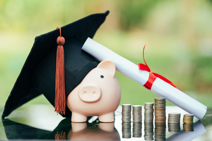 A diploma roll propped against piggy bank wearing a mortarboard with several small stacks of coins next to it 
