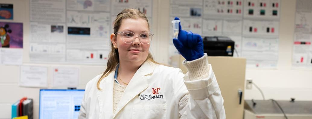 Hana Schwierling wears a white lab coat and latex gloves while holding a test tube up and examining inside