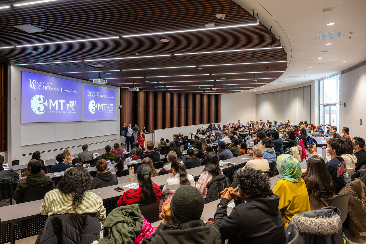 Audience members fill a large University of Cincinnati lecture hall during a Three Minute Thesis (3MT) event. Two presentation screens at the front display the 3MT logo, while attendees sit in tiered seating facing the stage area and presenters. The modern auditorium features wood-paneled walls, curved architecture, and overhead linear lighting.