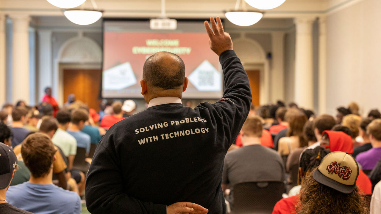 behind view of man standing with three fingers raised in a shirt that says solving problems with technology