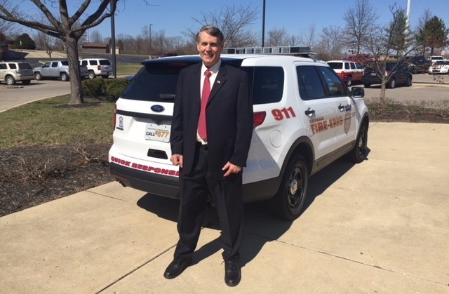 A middle-aged man in a dark suit and red tie stands outside on a sidewalk next to a sport utility vehicle that has "911" "Fire EMS" "Quick response team" decal on it