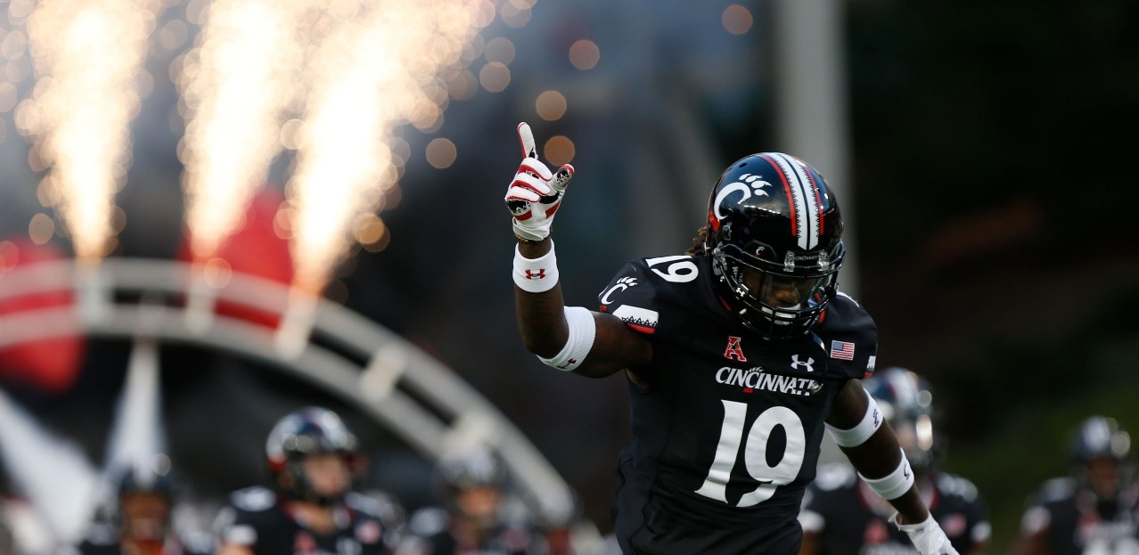 UC football player runs on to the field, holding up one finger while pyrotechnics light up the sidelines. 