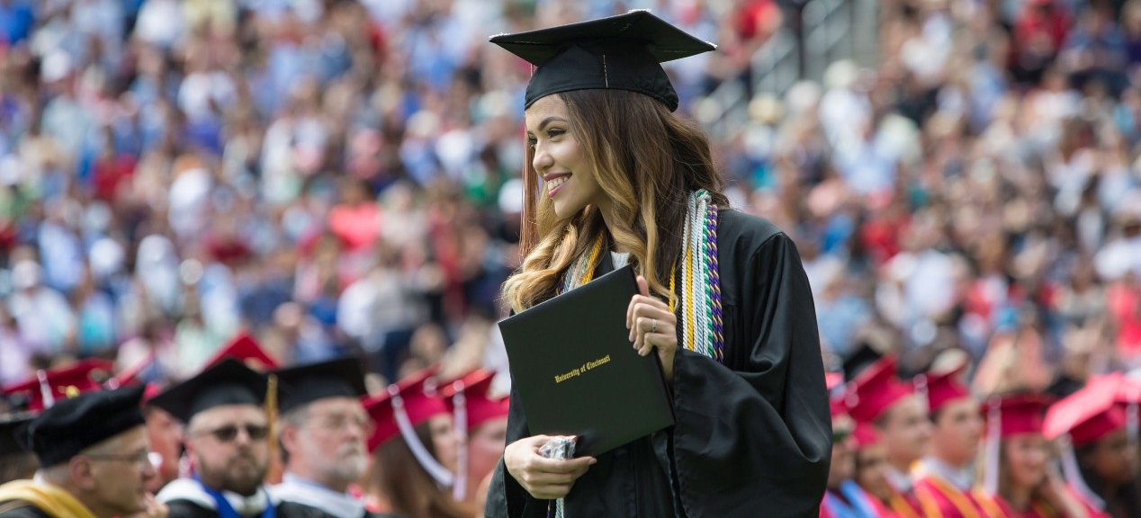 A UC student smiles to the crowd and proudly holds up her diploma at commencemnt. 