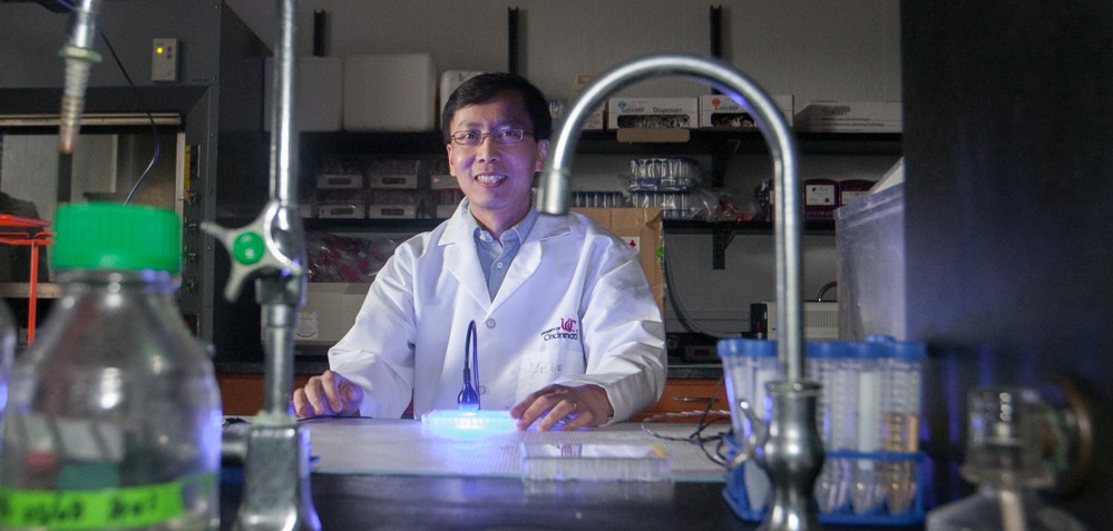 UC chemist Peng Zhang in a labcoat stands  in the dark at his lab bench illuminated by the glow of a blue LED. 