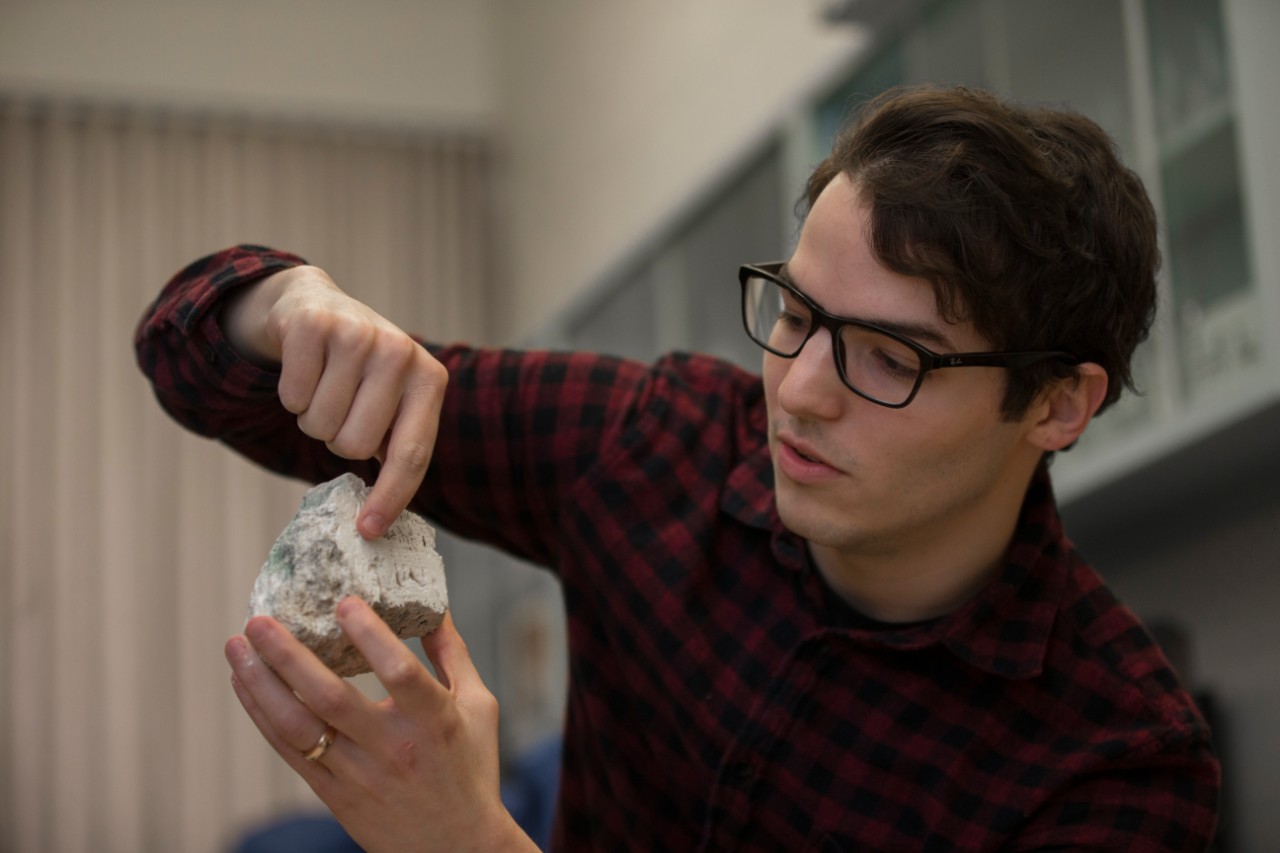 UC student Andrew Gangidine holds up and gestures to a layered rock.