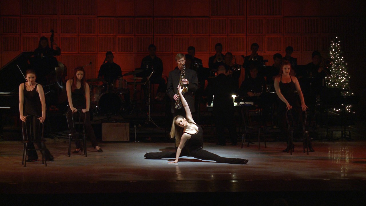 Dancers on stage with chairs, one in the spotlight doing the splits with musicians behind her
