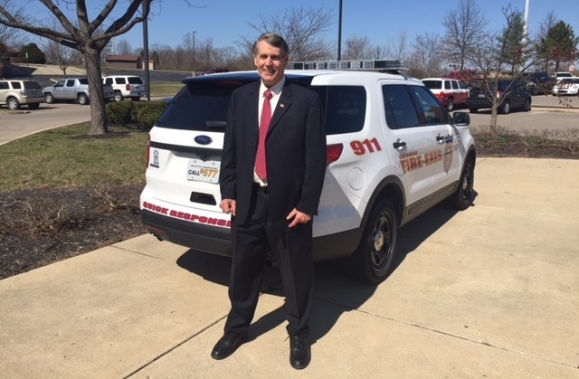 Man in dark suit, white shirt and red tie stands outside in front of an sport utility vehicle that has decals that read "911" and "Quick response team" and "Fire EMS"